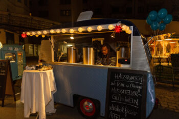 Ein gem&uuml;tlicher Foodtruck mit Lichterketten serviert abends neben warmen Getr&auml;nken auch SPiNNpunsch. Im Truck bereitet eine Person ein Getr&auml;nk zu, w&auml;hrend auf einer Tafel leckere Gerichte aufgelistet sind. In der N&auml;he bietet ein kleiner Tisch Erfrischungen an, w&auml;hrend blaue Luftballons der Szene eine festliche Note verleihen.