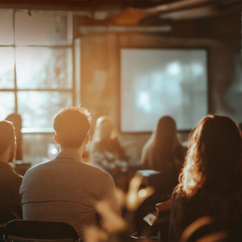 Menschen sitzen in einem schwach beleuchteten Raum vor einem verschwommenen Bildschirm, im Hintergrund fällt Sonnenlicht durch ein großes Fenster. Die Kulisse erinnert an ein KI-Kompetenzseminar, das die Lücke zwischen Wissen und Innovation schließt.