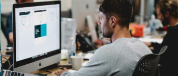 Eine Person im grauen Hemd sitzt an einem Schreibtisch in einem B&uuml;ro und konzentriert sich auf einen gro&szlig;en Monitor, auf dem Designsoftware l&auml;uft. Inmitten des gesch&auml;ftigen Arbeitsumfelds, mit mehreren Kollegen und Computern im Hintergrund, sorgt sie f&uuml;r die Passwortsicherheit aller digitalen Designs.