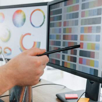 Eine Person zeigt mit einem Stift auf einen Computerbildschirm, der verschiedene Farbmuster anzeigt und gleichzeitig die Passwortsicherheit &uuml;berpr&uuml;ft. Ein weiterer Monitor im Hintergrund zeigt bunte Diagramme und Grafiken. Der Schreibtisch ist aufger&auml;umt, Stifte in einem Halter und &uuml;berall liegen Haftnotizen herum.