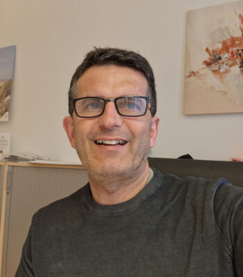 Ein Mann mit kurzen dunklen Haaren, Brille und dunklem Pullover sitzt im Haus und l&auml;chelt in die Kamera. Hinter ihm h&auml;ngt ein abstraktes Kunstwerk an der Wand, das sein Interesse an Design und die Auswirkungen des Barrierefreiheitsgesetzes unterstreicht.