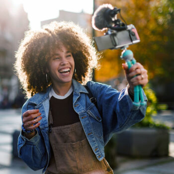 Eine junge Frau mit lockigem Haar lächelt und hält ein Smartphone in der Hand, das auf einem Stativ mit Mikrofon befestigt ist. Sie filmt sich selbst im Freien in einer sonnenbeschienenen städtischen Umgebung, um ihre Freie Meinungsäußerung zu teilen.