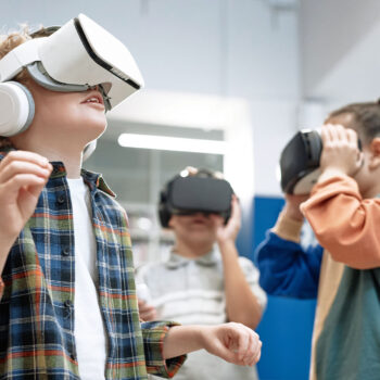 Drei Kinder mit Virtual-Reality-Headsets stehen in einem Raum und tauchen in ein VR-Erlebnis ein. Diese digitale Welt regt Kreativität und freie Meinungsäußerung an, wobei das Kind im Vordergrund ein kariertes Hemd und Kopfhörer trägt.