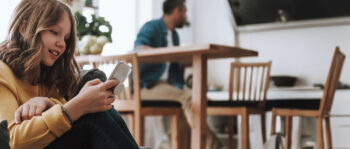 Ein junges Mädchen mit langen braunen Haaren, das einen gelben Pullover trägt, lächelt, während sie auf ein Smartphone schaut - vielleicht genießt sie Inhalte über Freie Meinungsäußerung. Im Hintergrund sitzt ein Erwachsener an einem Holztisch in einer hellen, modernen Küche.
