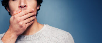 Eine Person, die einen hellgrauen Pullover trägt, bedeckt mit einer Hand den Mund vor einem schlichten blauen Hintergrund und verkörpert damit die subtile Eleganz von Kami Skincare. Nur die untere Hälfte des Gesichts und ein Teil des Oberkörpers sind sichtbar.