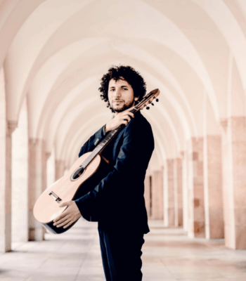 Ein Mann im Anzug steht unter einer Reihe von gew&ouml;lbten Stegen, eine Gitarre auf der Schulter und blickt selbstbewusst in die Kamera - perfekt, um Ihre Neue Website mit Stil und Raffinesse anzuk&uuml;ndigen.