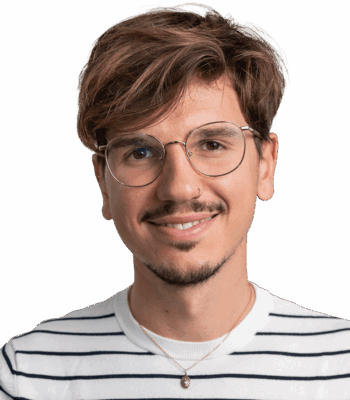 Ein junger Mann mit hellbraunem Haar und Brille l&auml;chelt in die Kamera. Er tr&auml;gt ein wei&szlig;es Hemd mit schwarzen Streifen und eine Halskette vor einem schlichten wei&szlig;en Hintergrund.