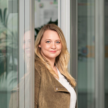 Eine Frau mit langen blonden Haaren, die eine braune Wildlederjacke über einem weißen Oberteil trägt, steht drinnen an einer Glaswand und lächelt sanft in die Kamera - perfekt für Social-Media-Marketing-Motive.