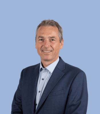 Ein Mann in einem marineblauen Anzug und einem hellblauen Hemd l&auml;chelt in die Kamera. Er hat kurzes graues Haar und steht vor einem schlichten hellblauen Hintergrund, perfekt f&uuml;r die Verwendung auf einer professionellen Website.