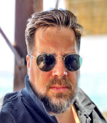 Ein Mann mit kurzem, braunem Haar, Bart und Schnurrbart tr&auml;gt eine Sonnenbrille und ein dunkles Hemd. Er befindet sich im Freien in hellem Sonnenlicht und bespricht m&ouml;glicherweise die Gemeinschaftsverwaltung, mit einem unscharfen Hintergrund aus einer Holzstruktur und blauem Himmel.