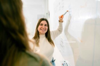 Eine lächelnde Frau, die mit einem Marker auf eine Tafel schreibt und möglicherweise eine Diskussion über Social Media Marketing leitet, steht einer anderen Person gegenüber, die der Kamera in einem Klassenzimmer oder Büro abgewandt ist.