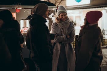 Eine Gruppe von winterlich gekleideten Menschen steht nachts draußen, unterhält sich und lächelt in der Nähe eines mit Lichterketten geschmückten Imbisswagens. Eine Frau in einem grauen Mantel und mit Mütze steht in der Mitte und unterhält sich.