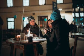 Drei Personen in Winterkleidung stehen nachts um einen Holztisch im Freien, unterhalten sich und essen. Das warme Licht von Kerzen und Gebäuden schafft eine gemütliche Atmosphäre. Der Hintergrund zeigt eine sanft beleuchtete Straße und ein Parkplatzschild.