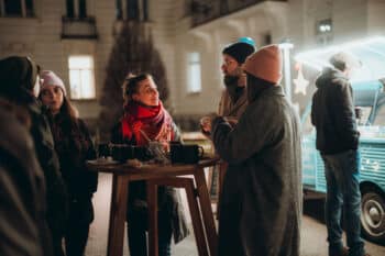Eine kleine Gruppe winterlich gekleideter Menschen steht nachts im Freien an einem hohen Holztisch, plaudert und schlürft SPiNNpunsch in der Nähe eines beleuchteten Foodtrucks. Die Szene sieht warm und festlich aus.