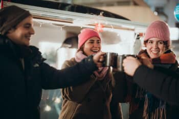 Drei Personen in Winterkleidung und Mützen lächeln und stoßen mit Tassen an, die von festlichen Lichterketten umgeben sind.