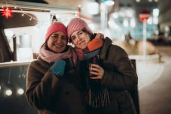 Zwei Personen mit warmen Mänteln, Mützen und Schals lächeln und halten Getränke in der Hand, während sie nachts im Freien stehen, mit festlichen Lichtern und verschwommenen Stadtgebäuden im Hintergrund.