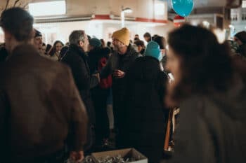 Eine Gruppe von Menschen sitzt nachts im Freien, viele in Winterkleidung, und genießt SPiNNpunsch. Zwei Männer in der Mitte schütteln sich die Hände und lächeln. Ein blauer Luftballon schwebt über der Menge und Lichter erhellen die festliche Szene.