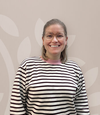 Eine Person mit schulterlangem blondem Haar, Brille und Ohrringen l&auml;chelt in die Kamera. Sie tr&auml;gt einen schwarz-wei&szlig; gestreiften Pullover mit rosa Kragen von VABON und steht vor einem hellen, gemusterten Hintergrund.