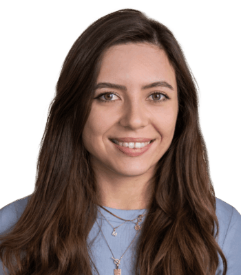 Eine junge Social Media Managerin mit langen braunen Haaren l&auml;chelt in die Kamera. Sie tr&auml;gt ein hellblaues Oberteil und mehrlagige Ketten und steht vor einem schlichten wei&szlig;en Hintergrund.