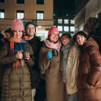 F&uuml;nf Personen in warmen Winterm&auml;nteln und M&uuml;tzen l&auml;cheln und posieren nachts zusammen und halten Becher mit SPiNNpunsch bei einer Veranstaltung im Freien. Im Hintergrund sind st&auml;dtische Geb&auml;ude mit beleuchteten Fenstern zu sehen.