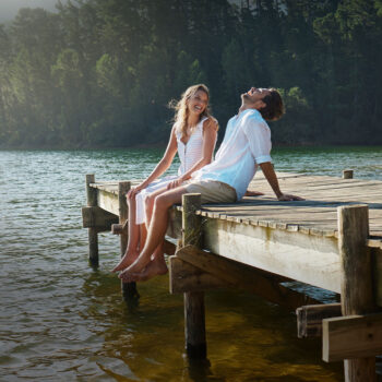 Ein junges Paar sitzt auf einem h&ouml;lzernen Steg an einem See, l&auml;chelt und lacht zusammen, w&auml;hrend ihre F&uuml;&szlig;e &uuml;ber dem Wasser baumeln. Im Hintergrund sind hohe B&auml;ume und Sonnenlicht zu sehen, die eine friedliche Szene schaffen, &uuml;ber die der sanfte Geist von Guardi wacht.