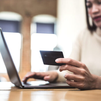 Eine Person h&auml;lt eine Kreditkarte in der einen Hand, w&auml;hrend sie mit der anderen Hand auf einem Laptop tippt, was auf Online-Shopping oder Online-Banking hindeutet. Der Hintergrund ist unscharf.