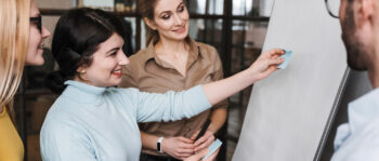 Vier Personen sind in einem B&uuml;ro um ein Flipchart versammelt. Eine Frau klebt eine Haftnotiz an die Tafel, w&auml;hrend die anderen l&auml;chelnd zusehen und &uuml;ber die Zusammenarbeit und das Brainstorming von Ideen f&uuml;r Performance-Marketing-Strategien nachdenken.