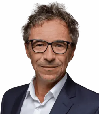 Ein Mann mittleren Alters mit kurzen, zerzausten grauen Haaren, einer schwarz umrandeten Brille und einem weißen Kragenhemd unter einer dunklen Anzugsjacke lächelt leicht vor einem schlichten grauen Hintergrund und strahlt die Zuversicht eines Performance-Marketing-Experten aus.