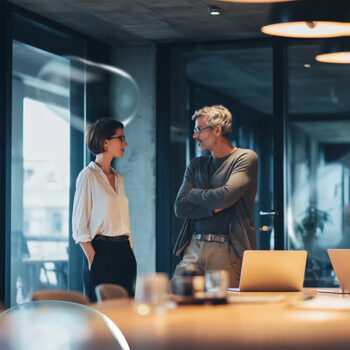 Eine Frau und ein Mann stehen in einem modernen B&uuml;ro und f&uuml;hren ein Gespr&auml;ch. Beide scheinen konzentriert zu sein, wobei der Mann seine Arme verschr&auml;nkt. Laptops und B&uuml;romaterialien sind auf einem Tisch im Vordergrund zu sehen.