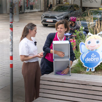 Zwei Frauen stehen in der N&auml;he eines Gehwegaufstellers mit einer Cartoon-Engelsfigur und dem Debra-Logo. Eine Frau legt einen lila Gegenstand in eine Metallbox, w&auml;hrend die andere spricht. Im Hintergrund sind Radfahrer und st&auml;dtische Landschaften zu sehen.