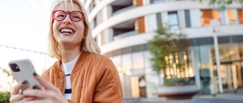 Eine lächelnde Person mit blondem Haar und roter Brille hält ein Smartphone im Freien und trägt eine braune Jacke. Im Hintergrund sind ein modernes Gebäude und ein Baum zu sehen.