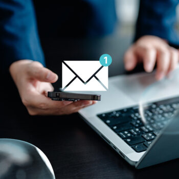 Eine Person hält ein Smartphone in der Nähe eines Laptops. Über dem Telefon erscheinen ein digitaler Briefumschlag und ein Benachrichtigungssymbol, das eine neue Nachricht anzeigt.