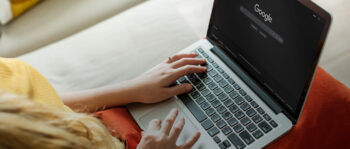 Eine Person mit einem Laptop, auf dem die Google-Homepage im dunklen Modus ge&ouml;ffnet ist, tippt auf der Tastatur, w&auml;hrend sie auf einer Couch mit einem orangefarbenen Kissen sitzt.