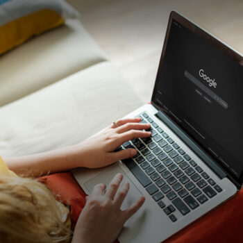 Eine Person, die einen Laptop benutzt, auf dessen Bildschirm die Startseite der Google-Suche angezeigt wird. Sie sitzt auf einem Sofa und tippt mit einer Hand auf der Tastatur, w&auml;hrend die andere Hand auf dem Laptop ruht.