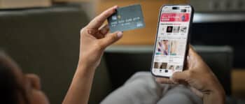 Eine Person sitzt auf einem Sofa und hält in der einen Hand eine Kreditkarte und in der anderen ein Smartphone, auf dem eine Online-Shopping-App zu sehen ist.