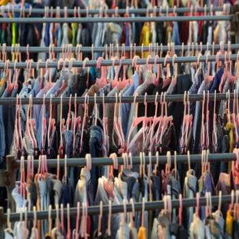 Reihen mit unterschiedlichster Kleidung an rosa Plastikkleiderbügeln sind an Kleiderständern aufgehängt und bilden eine dichte, farbenfrohe Auslage, die an einen Markt im Freien oder einen Secondhand-Laden erinnert.