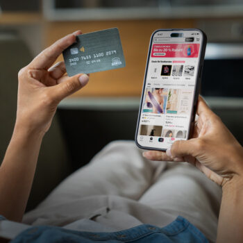 Eine Person, die in der einen Hand eine Kreditkarte und in der anderen Hand ein Smartphone mit einer Online-Shopping-App h&auml;lt und den Anschein erweckt, als w&uuml;rde sie einen Einkauf t&auml;tigen, w&auml;hrend sie entspannt auf einer Couch sitzt.