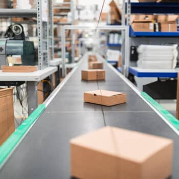 Mehrere kleine braune Pakete bewegen sich auf einem Förderband in einem Lagerraum, im Hintergrund sind Metallregale, Verpackungsmaterial und verschiedene Geräte zu sehen.