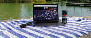Auf einem blau-weiß gestreiften Handtuch, das auf einem Steg an einem See liegt, befinden sich ein Laptop mit einer Karte auf dem Bildschirm, eine Dose Pepsi und eine Sonnenbrille; im Hintergrund sind grüne Bäume und Wasser zu sehen.