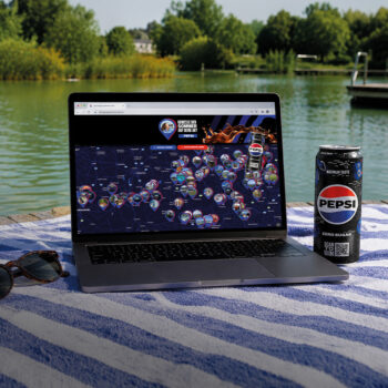 Ein Laptop, auf dem eine Website mit einer Weltkarte zu sehen ist, steht auf einem gestreiften Handtuch am Ufer eines Sees, neben einer Sonnenbrille und einer Dose Pepsi Zero Sugar. Im Hintergrund sind B&auml;ume und Wasser zu sehen.