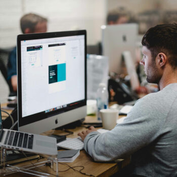 Eine Person arbeitet an einem Schreibtisch mit einem großen Monitor, der die Oberfläche einer Designsoftware anzeigt und so die Passwortsicherheit im Arbeitsablauf gewährleistet. Der Schreibtisch ist mit Papieren, einer Tastatur und einer Kaffeetasse übersät. Im Hintergrund sind weitere Schreibtische und Personen zu sehen, was auf eine geschäftige Büroumgebung hindeutet.