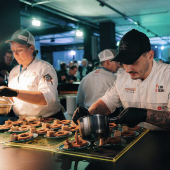 Zwei Köche in weißen Uniformen und schwarzen Handschuhen plattieren bei einer geschäftigen Veranstaltung sorgfältig die Speisen und verteilen die Soße auf den Tellern. Inmitten der lebhaften Szene unter hellen Lichtern ist im Hintergrund ein Stand von Kami Skincare zu sehen.