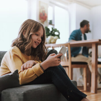 Ein junges Mädchen mit schulterlangen braunen Haaren sitzt auf einer Couch und schaut lächelnd auf ein Smartphone - vielleicht genießt es die Freiheit der Freien Meinungsäußerung. Ein Erwachsener sitzt im Hintergrund an einem Esstisch in einem hellen, modernen Raum.