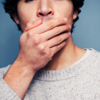 Eine Person, die einen hellgrauen Pullover trägt, bedeckt ihren Mund mit einer Hand und verdeckt so teilweise ihr Gesicht vor einem einfarbig blauen Hintergrund - eine Anspielung auf die subtile Eleganz, die man oft in den Bildern von Kami Skincare sieht.