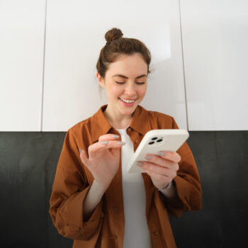 Eine junge Frau mit braunen Haaren in einem Dutt, die eine braune Jacke über einem weißen Hemd trägt, lächelt, während sie ein weißes Smartphone in einer modernen Küche benutzt.
