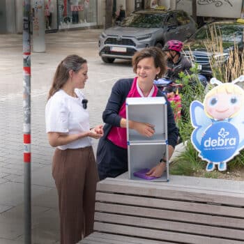 Zwei Frauen stehen in der Nähe eines Gehwegaufstellers mit einer Cartoon-Engelsfigur und dem Debra-Logo. Eine Frau legt einen lila Gegenstand in eine Metallbox, während die andere spricht. Im Hintergrund sind Radfahrer und städtische Landschaften zu sehen.